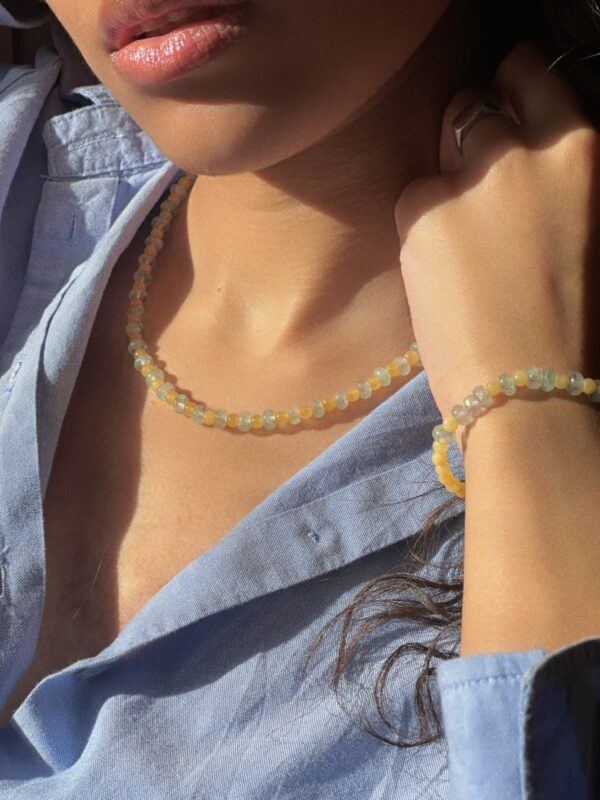 try-on shot of the hand-beaded Citrine Skies set of necklace and bracelet in butter yellow agate stones and tourmaline blue stones. A soft summer day, captured in stone. Inspired by the golden warmth of citrine sunsets and the tranquility of clear skies. Blends butter yellow and sky blue tones in a dreamy, refreshing palette. Handcrafted with natural jade and tourmaline gemstones and finished with 925 sterling silver clasp. Material: Natural jade stones - Length: 42 cm - 925 sterling silver clasp.