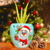 Christmas themed blue plant pot with Aloe Vera plant, pinecones, and holiday background. | اصيص زرع أزرق بتصميم الكريسماس يحتوي على نبات صبار، بجانب أكواز الصنوبر وخلفية احتفالية.