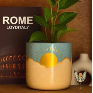 أصيص زرع فخار كبير بتصميم سماوي يظهر شمس ذهبية ساطعة فوق سحب بيضاء. | Large clay planter with a celestial design featuring a bright golden sun over white clouds.