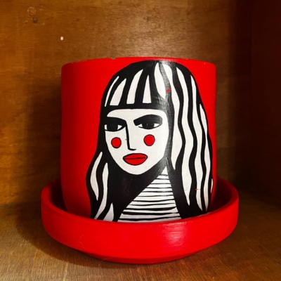 the Red Girl - Terracotta planter Pot | Only One Piece 2 أصيص فخاري أسطواني أحمر زاهٍ مرسوم يدوياً بتصميم جرافيكي لوجه امرأة بالأسود والأبيض. الأصيص يزرع به نبات الثعبان (Sansevieria)، وموضوع على لوح تقطيع خشبي، والخلفية مطبخ عصري بخزائن خضراء داكنة.| A vibrant red, cylindrical terracotta planter hand-painted with a striking graphic, pop-art style portrait of a woman in black and white. The pot holds a snake plant (Sansevieria) and is displayed in a sunny, modern kitchen with dark green cabinets.