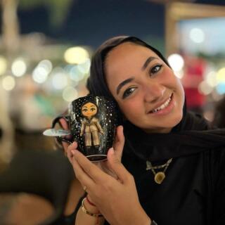 Handmade black mug featuring a custom polymer clay character of a woman wearing a hijab, designed to resemble the mug owner, held by a smiling woman.