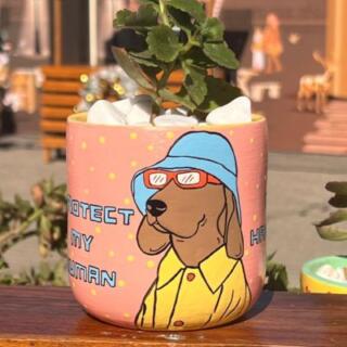 أصيص زرع فخار وردي مرسوم عليه كلب يرتدي قبعة ونظارات شمسية.| Pink clay planter featuring a hand-painted dog wearing a hat and sunglasses.