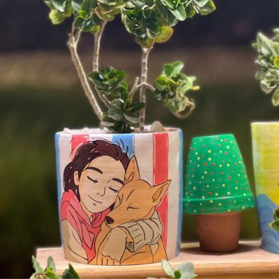 Girl & Dog Planter Pot 1 AR: أصيص زرع فخاري مرسوم يدوياً يظهر فتاة تحتضن كلبها مع خلفية مخططة. | Handmade Clay planter featuring a girl hugging her dog with a striped background.