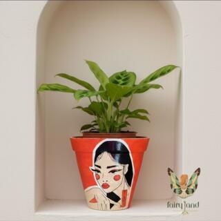 A close-up of a vibrant orange, hand-painted terracotta plant pot featuring an elegant, bold portrait of a woman with black hair, strong red makeup, and an Asian-style character earring. The pot sits on a wooden shelf in warm, natural light