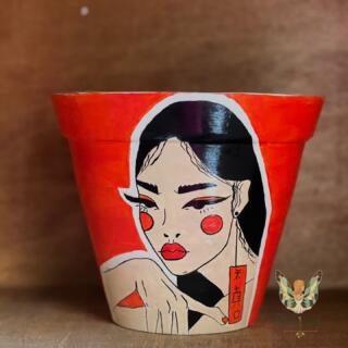 "Close-up of a vibrant orange, hand-painted terracotta planter featuring a bold, elegant portrait of a woman with Asian-inspired features and black hair. The pot is placed on a simple wooden shelf with soft natural lighting." (لقطة مقربة لأصيص فخاري برتقالي زاهي، مرسوم يدويًا بتصميم وجه امرأة أنيقة وجريئة بملامح آسيوية وشعر أسود. الأصيص موضوع على رف خشبي بسيط بإضاءة طبيعية ناعمة.)