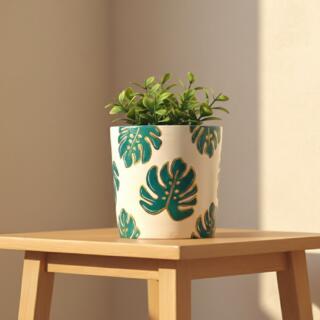 أصيص فخاري أسطواني مرسوم يدوياً بنمط متكرر لأوراق المونستيرا باللون الأخضر الداكن ومحدد باللون الذهبي. الأصيص موضوع في زاوية أنيقة بجوار كتاب وديكور عصري باللون البيج.| A cylindrical terracotta planter hand-painted with a repeating pattern of deep green Monstera leaves outlined in gold on a white/beige background. The pot is styled with books and modern arch decor in a warm, indoor setting.