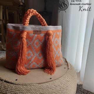 “Handmade crochet tote bag in terracotta orange and gray, featuring geometric patterns, braided handle, and long tassel details.