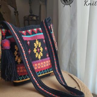 Handmade tapestry crochet crossbody bag in black with colorful geometric patterns, featuring a zipper closure, long strap, and side tassel.