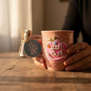 Pink handmade ceramic mug with white polka dots, decorated with a 3D clay badge saying “Eid Mubarak” in Arabic, embellished with small balloons, stars, and crystals