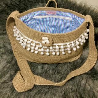 Handcrafted jute sling bag with white pom-pom trim, long woven strap, and soft checked fabric lining.