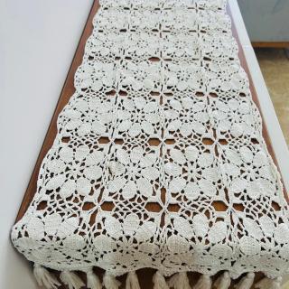 Handmade white crochet table runner with floral patterns and tassel edges displayed on a wooden table.