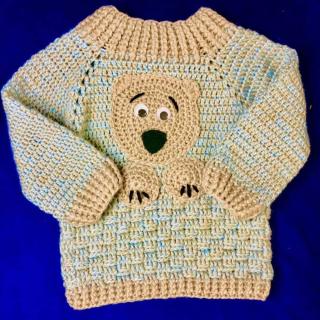 Handmade crochet baby set featuring a teddy bear sweater, matching knit hat with a pom-pom, and a textured scarf in soft beige and light blue colors.